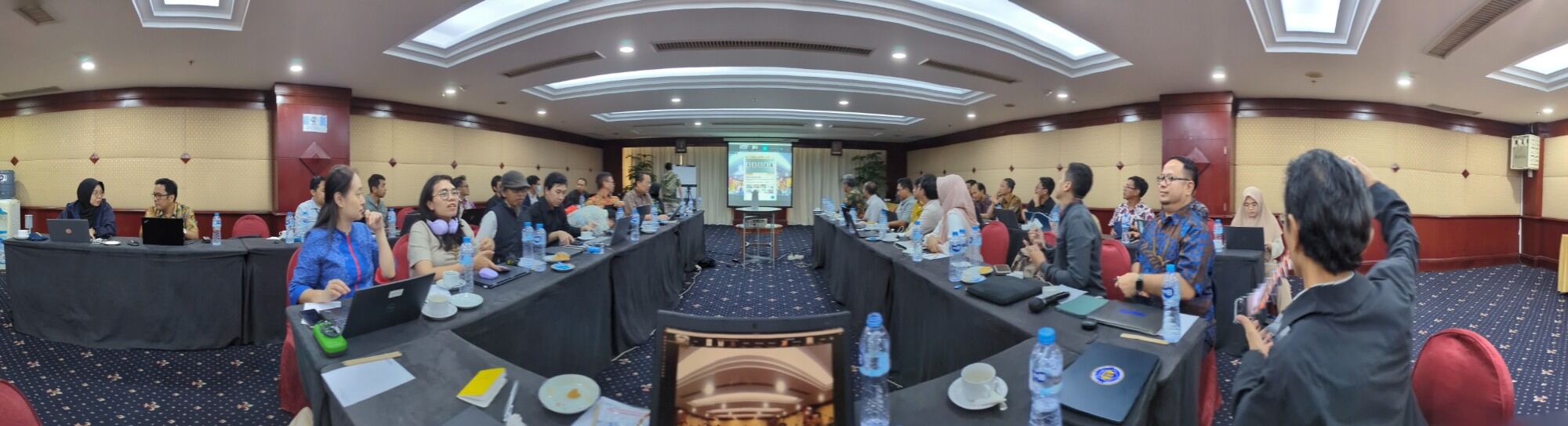 Panoramic view of a workshop room in Jakarta, with government participants seated at long tables facing a presentation screen.